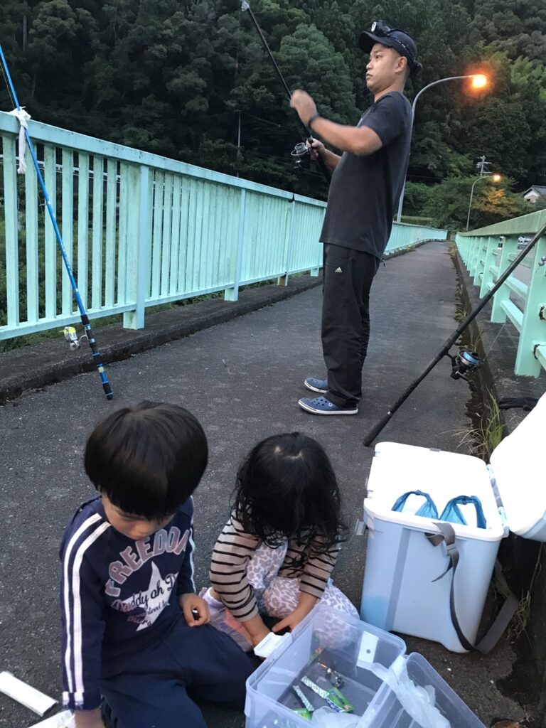 橋の上で男性が釣り竿を持ち、子ども2人が箱に入っているものを触っている